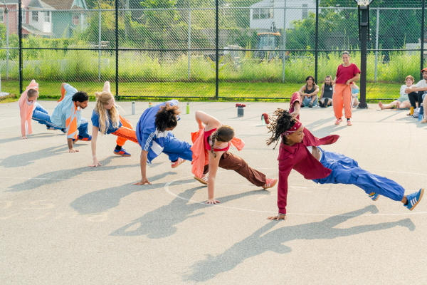 six people perform on a basketball court/community space, with an audience in the background